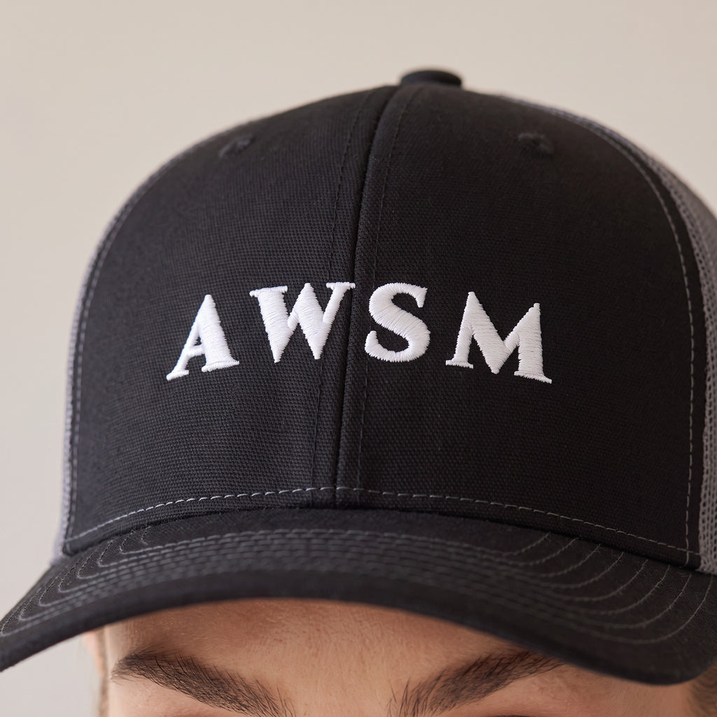 Minuit Trucker Cap — AWSM Maison in Onyx / Graphite, close-up of the embroidery on the AWSM house model